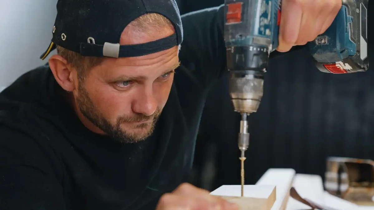 Homem usando uma furadeira elétrica para perfurar um pedaço de madeira, vestindo boné para trás e camiseta preta, concentrado no trabalho de carpintaria.