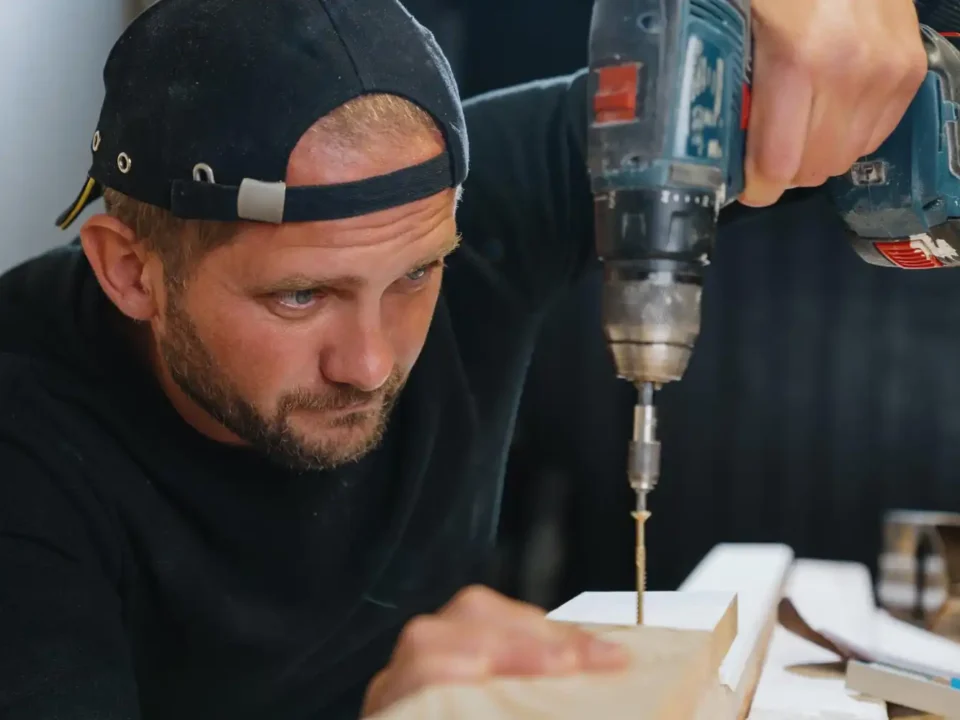 Homem usando uma furadeira elétrica para perfurar um pedaço de madeira, vestindo boné para trás e camiseta preta, concentrado no trabalho de carpintaria.