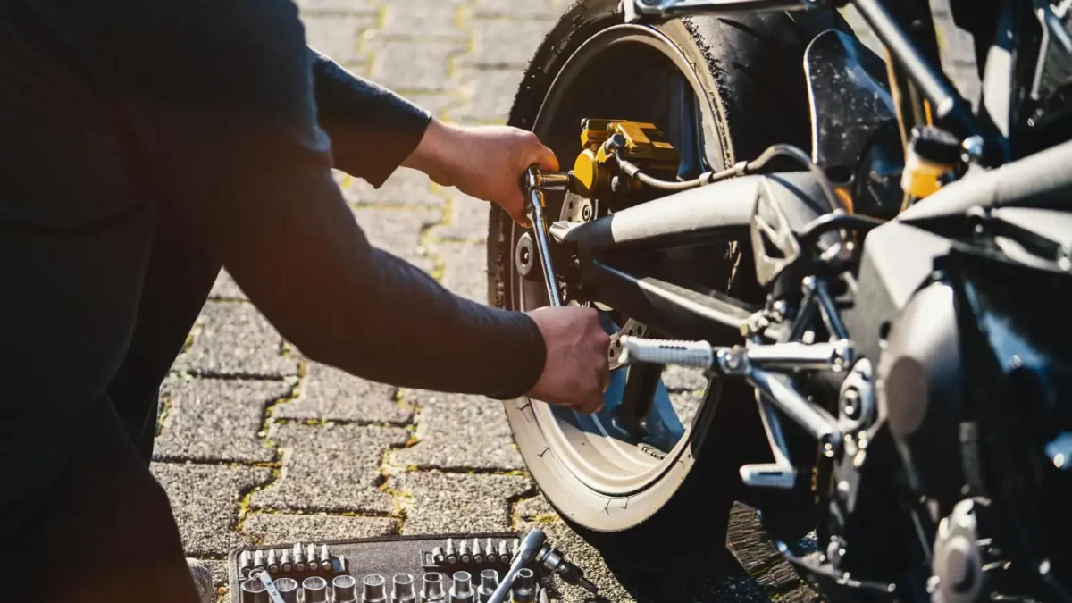 Mecânico utilizando um conjunto de soquetes para ajustar a roda traseira de uma motocicleta, com um kit de ferramentas visível no chão.