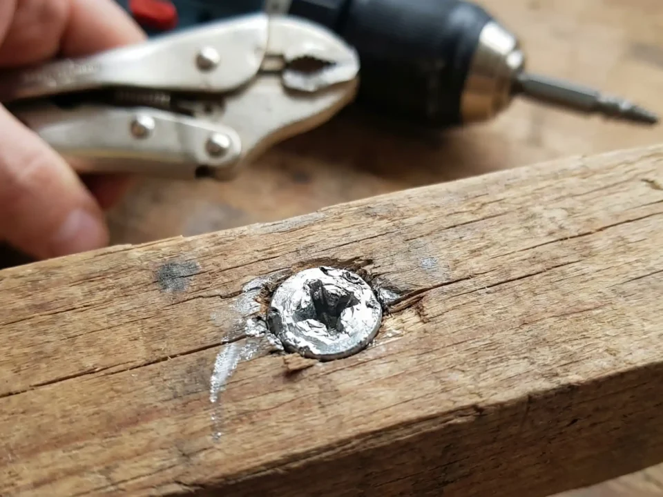 Close-up de um parafuso Phillips com a cabeça muito espanada cravado em um pedaço de madeira, com uma mão segurando um alicate de pressão e uma furadeira com extrator ao fundo.