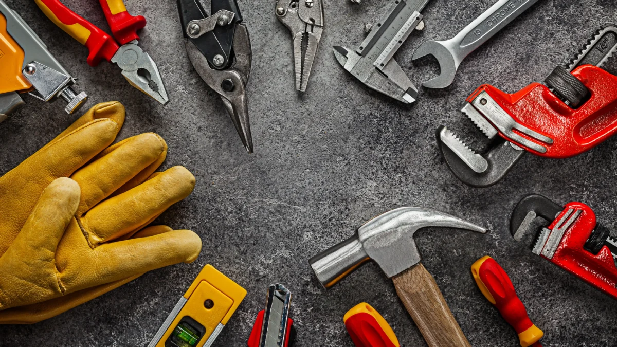 Ferramentas manuais organizadas sobre bancada escura, incluindo martelo, alicates, chave inglesa, chave de grifo, nível e luvas de proteção
