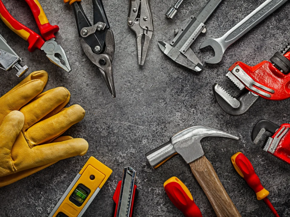 Ferramentas manuais organizadas sobre bancada escura, incluindo martelo, alicates, chave inglesa, chave de grifo, nível e luvas de proteção